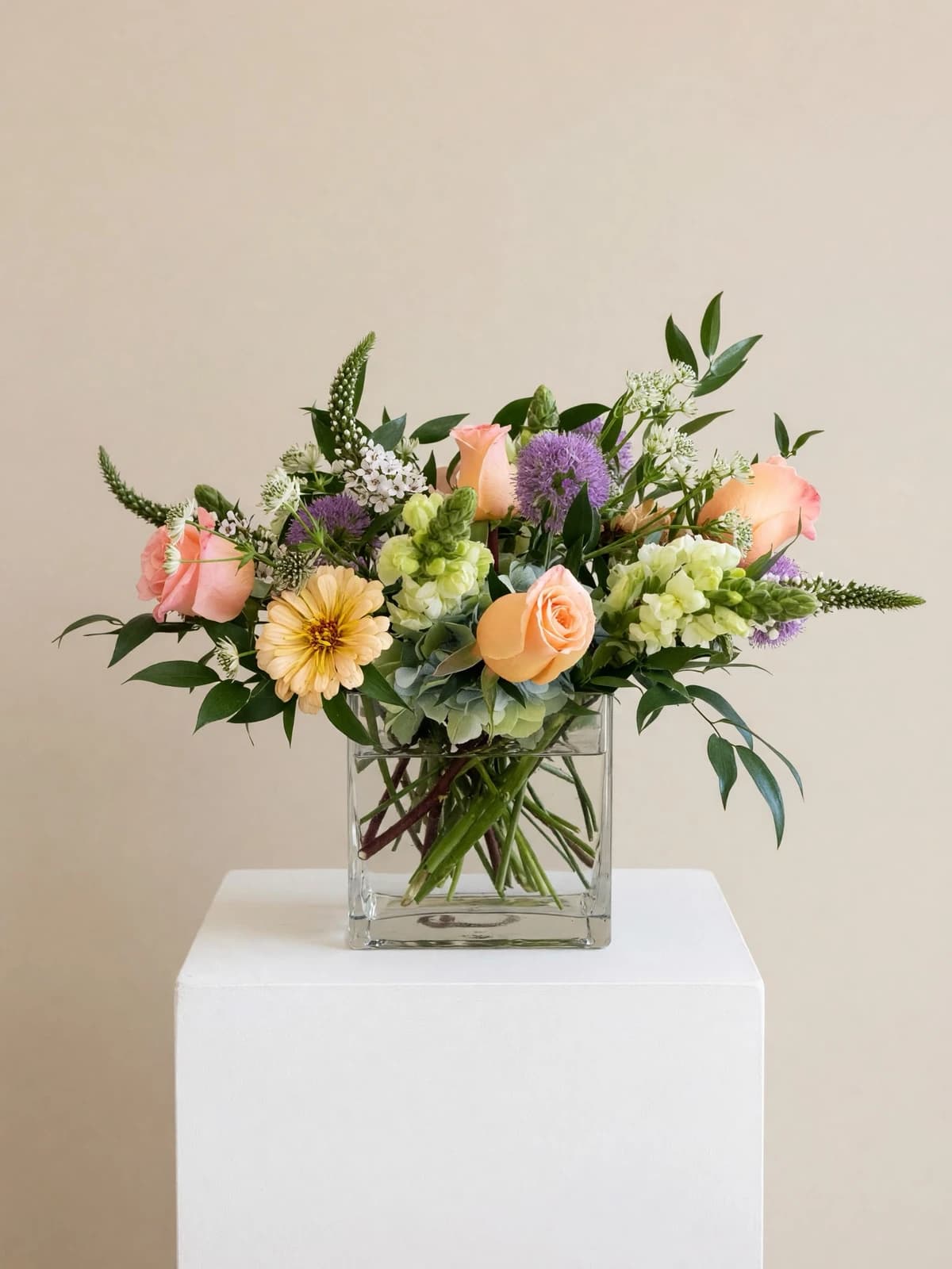 Soft peach, blush, and lavender tones blend with creamy whites and pale greens in Pastelo, an airy, garden-style design. Gentle spires and delicate little blooms add movement, while graceful greenery gives it a lush, natural shape. Arranged loosely in a clear square glass vase, it’s romantic and joyful.